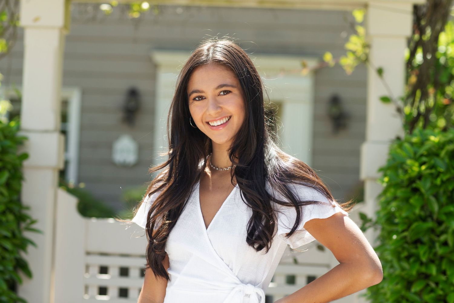 Smiling young woman in white short-sleeve wrap dress posing in front of a house with green shrubs.