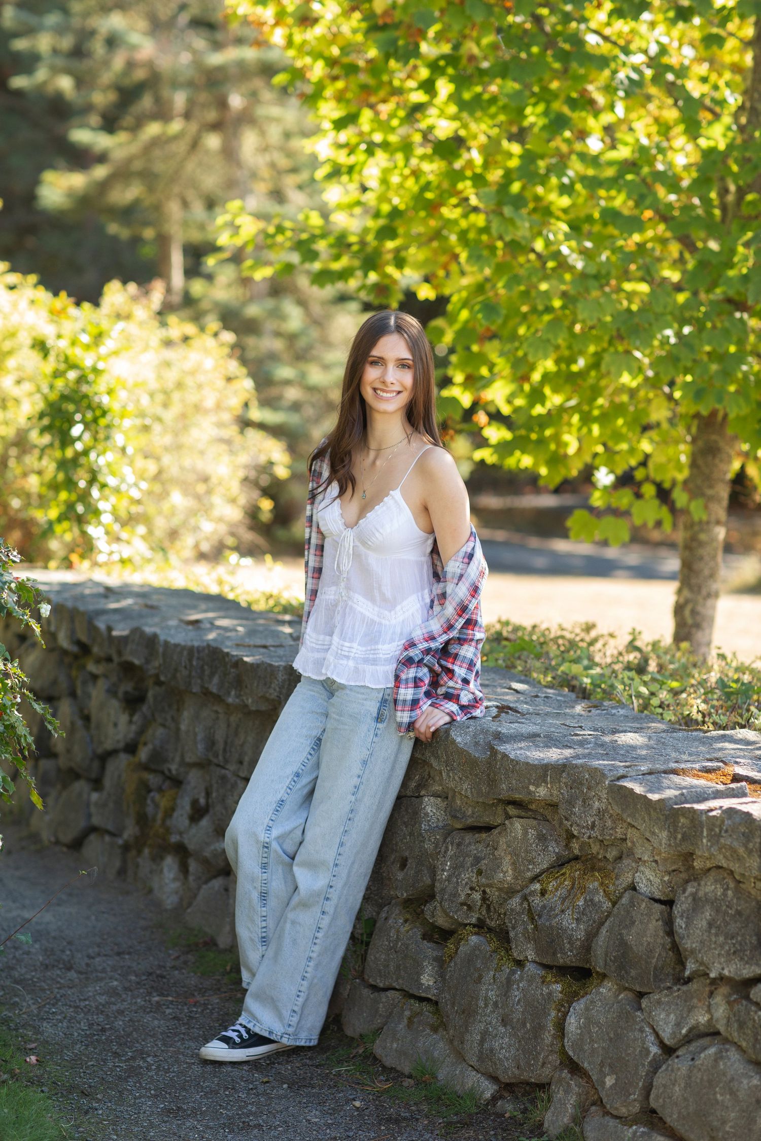 Smiling young woman leaning on stone wall in park wearing plaid shirt and wide-leg jeans.