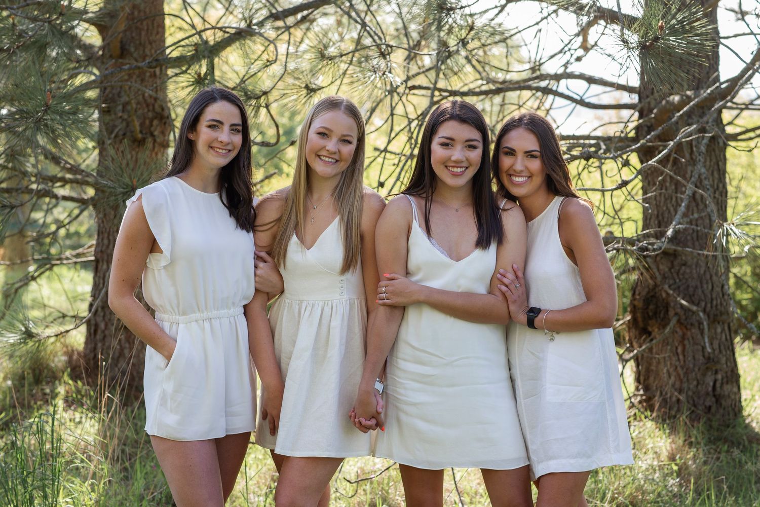 Four young women in white dresses smile together outdoors among pine trees in a bright, natural forest setting.