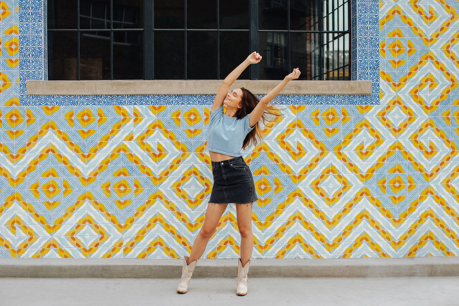 Young woman in blue top and black skirt dancing joyfully in front of a colorful geometric tiled wall.