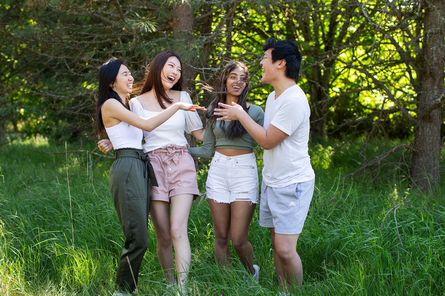 Four young friends laughing and toasting drinks together in a lush green park on a sunny day.