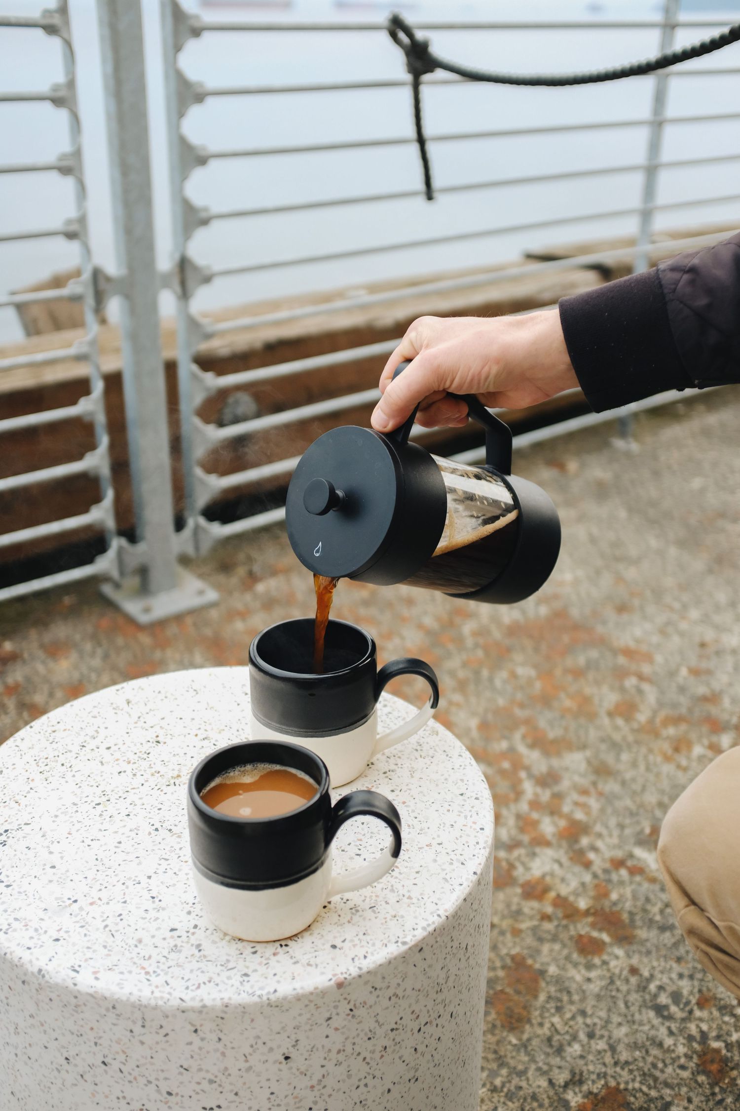 Hand pouring fresh coffee from a French press into black ceramic mugs on a white concrete table outdoors.