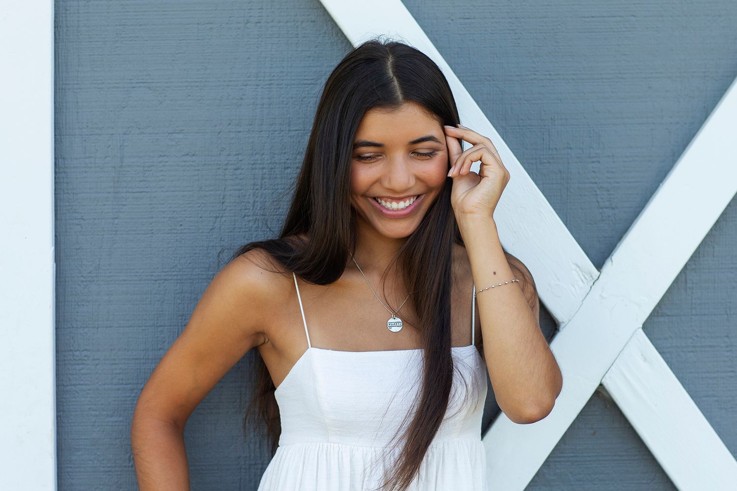 Smiling young woman in white sundress posing outdoors against a gray wall with white X detail.