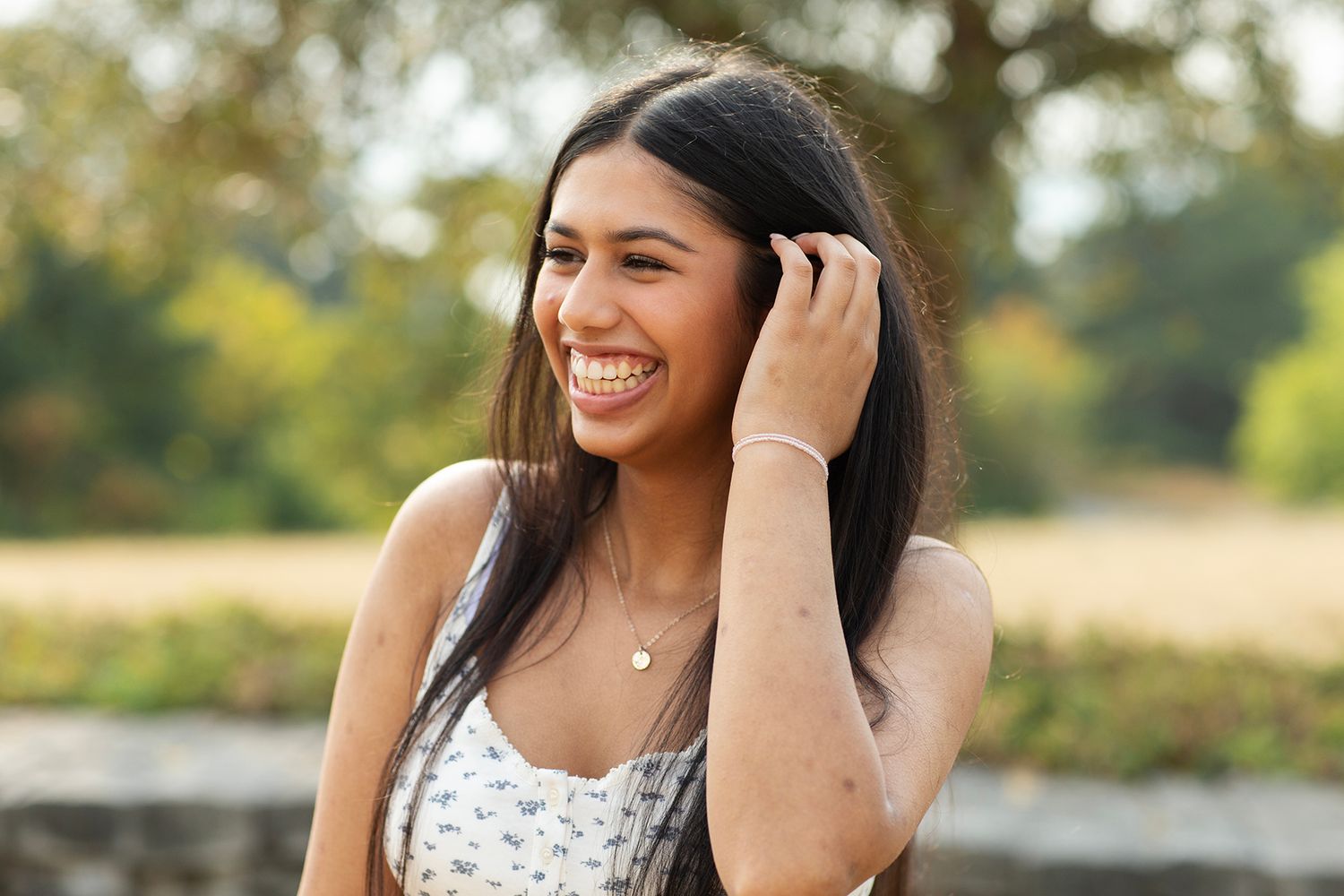 Laughing young woman with long dark hair in floral top outdoors with blurred green background.