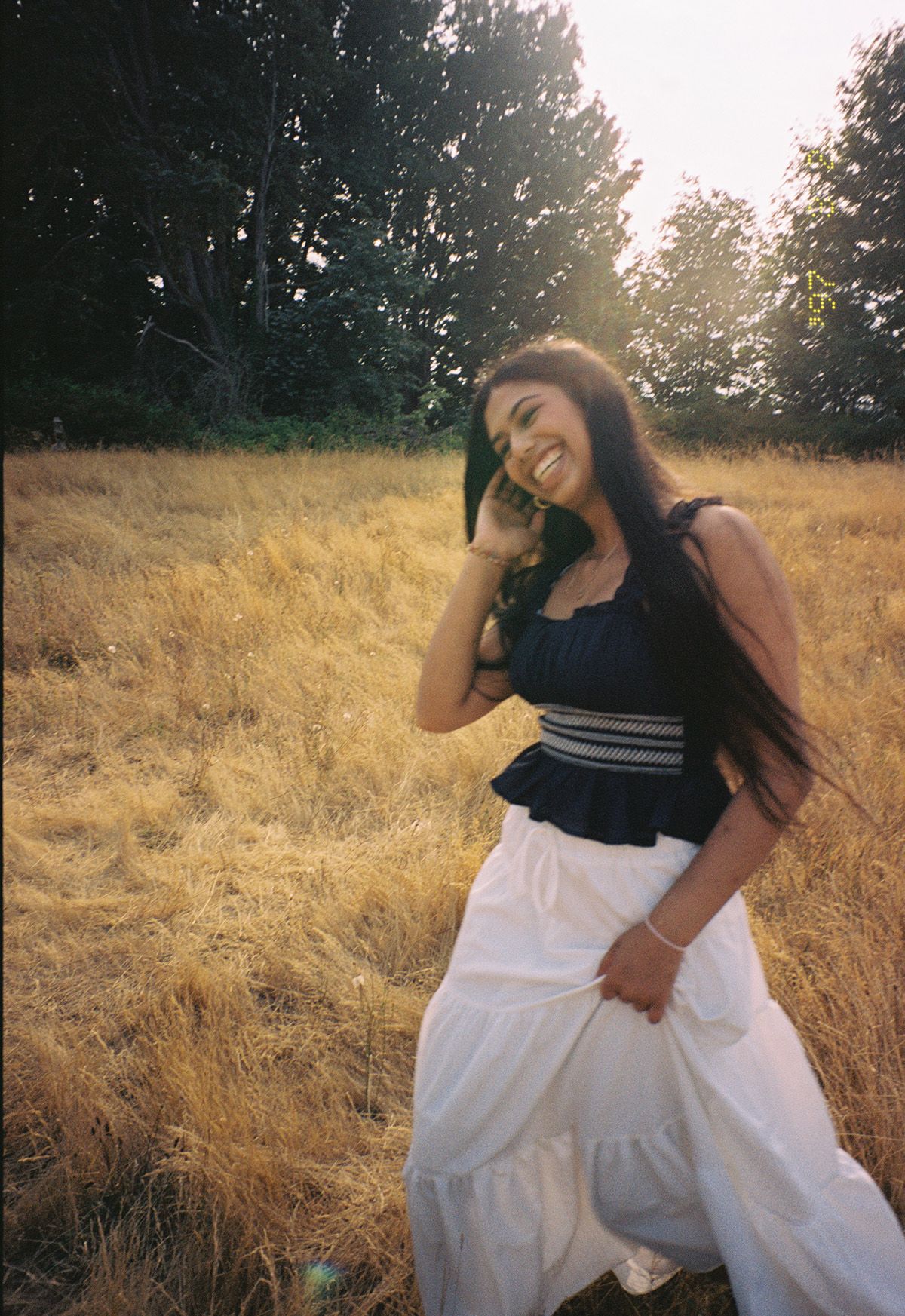 Laughing young woman in black top and white skirt posing in golden field at sunset.