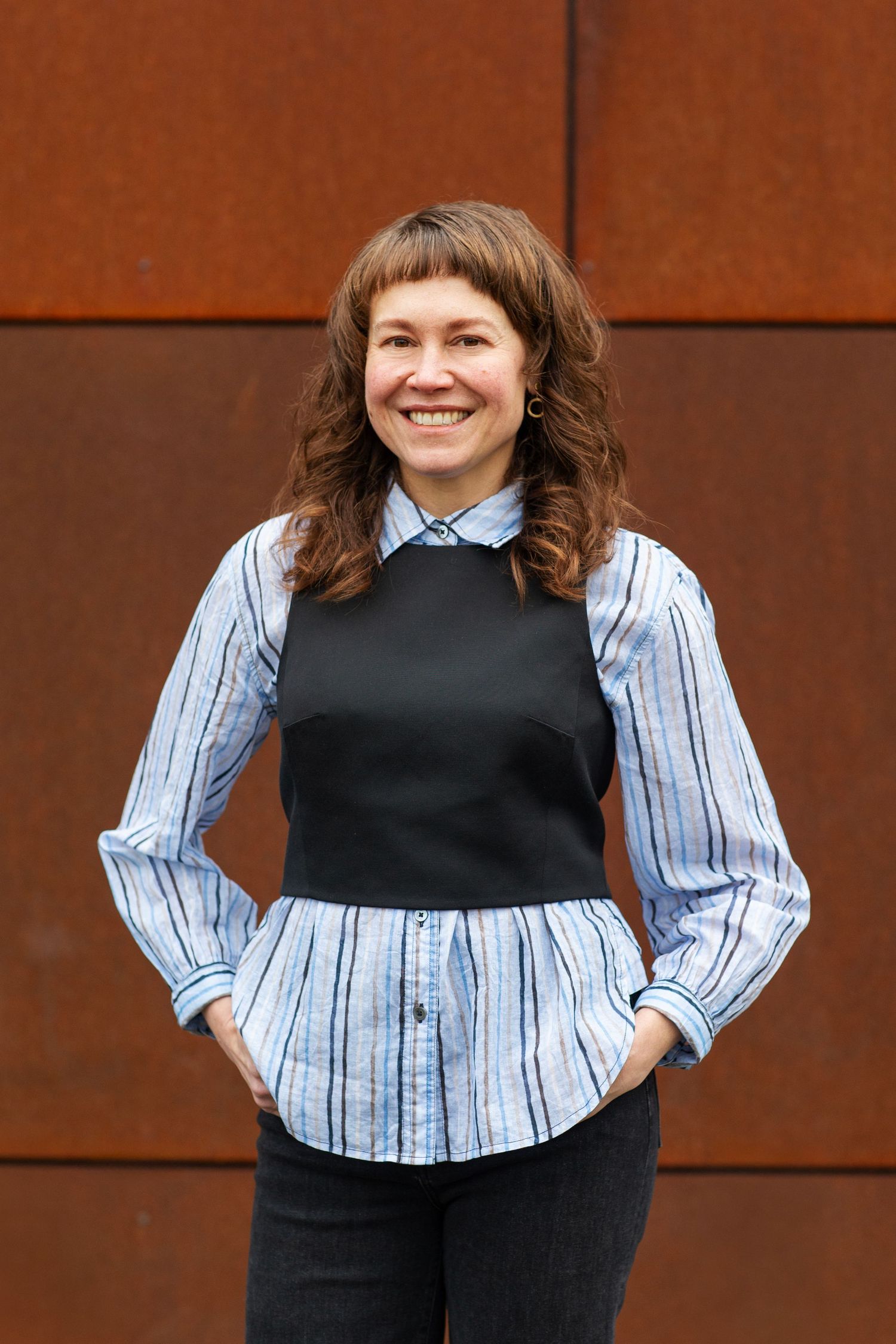 Cheerful woman in striped shirt and black vest posing against rustic metal wall.