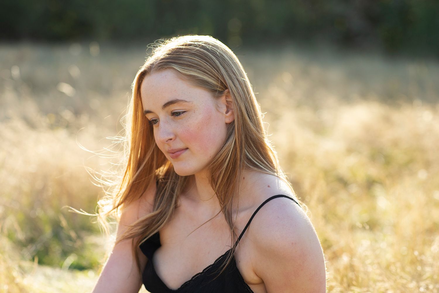 Young blonde woman with freckles sitting in golden sunlit field, wearing black top, hair blowing gently in breeze.