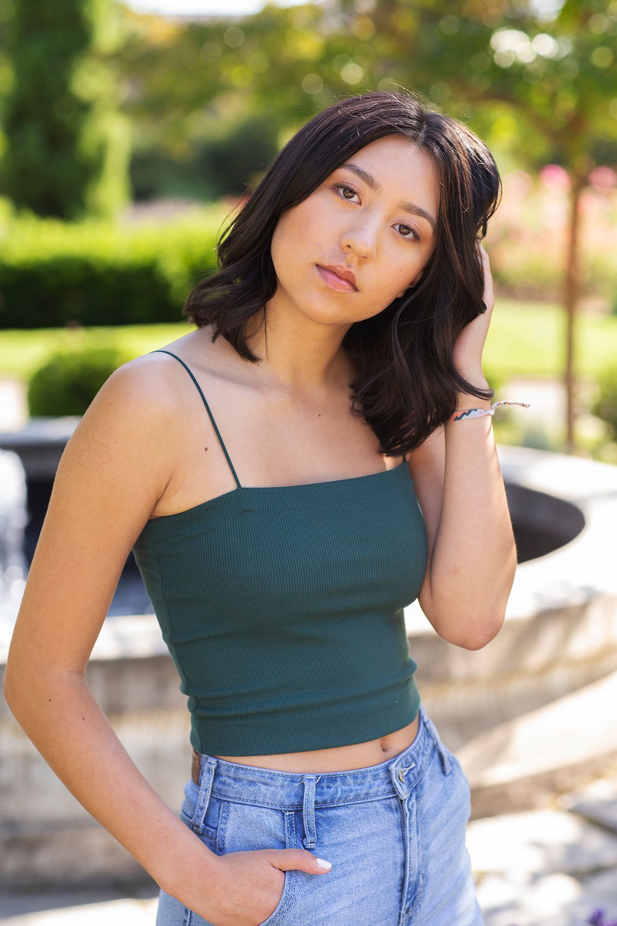Young Asian woman in green crop top and jeans poses outdoors in a sunny garden setting.