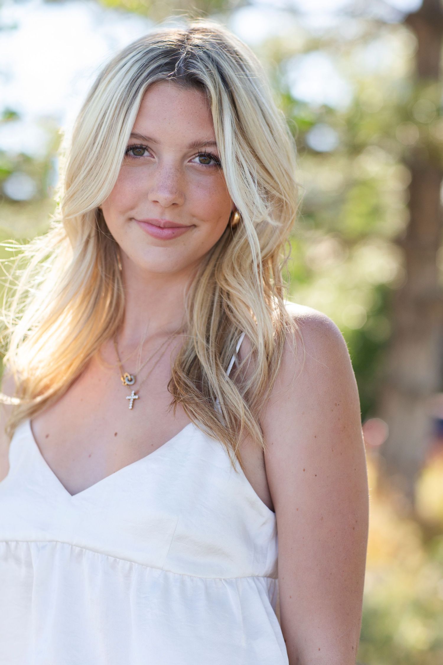 Close-up portrait of a smiling blonde woman wearing a white dress and delicate cross necklace outdoors.