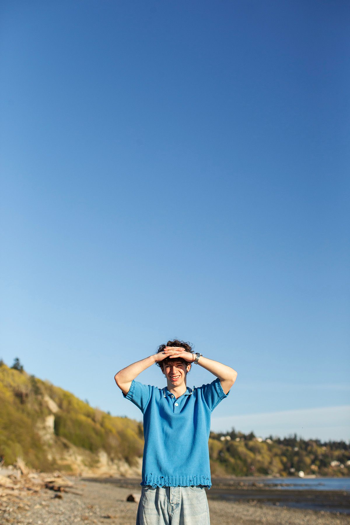 Young man in blue polo shirt stands on rocky beach, hands on head, smiling under clear blue sky.