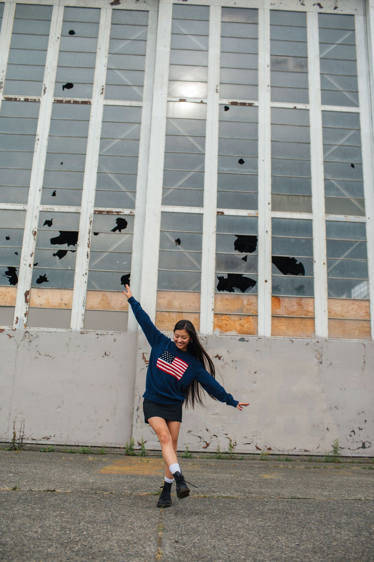 Young woman in navy American flag sweatshirt dancing playfully in front of industrial building with broken windows.