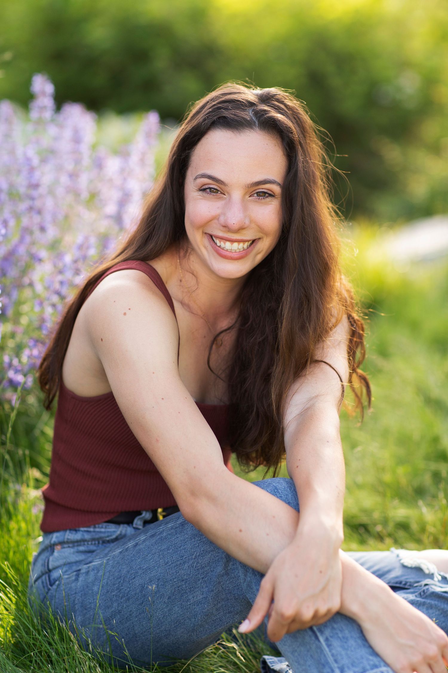 Smiling young woman with long brown hair sitting on grass near purple wildflowers in a red tank top and jeans.