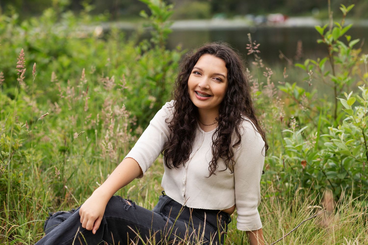 Young woman in cream sweater sitting in tall grass meadow near a peaceful lakeside.