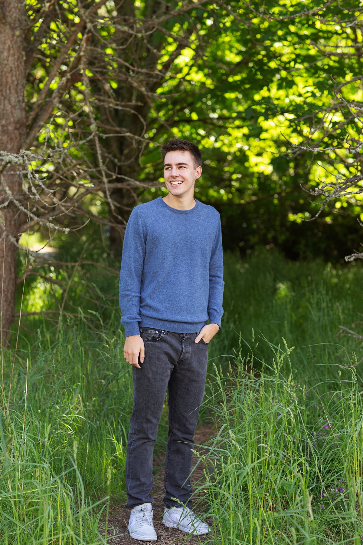 Smiling young man in blue sweater and gray jeans standing outdoors among lush green trees and tall grass.