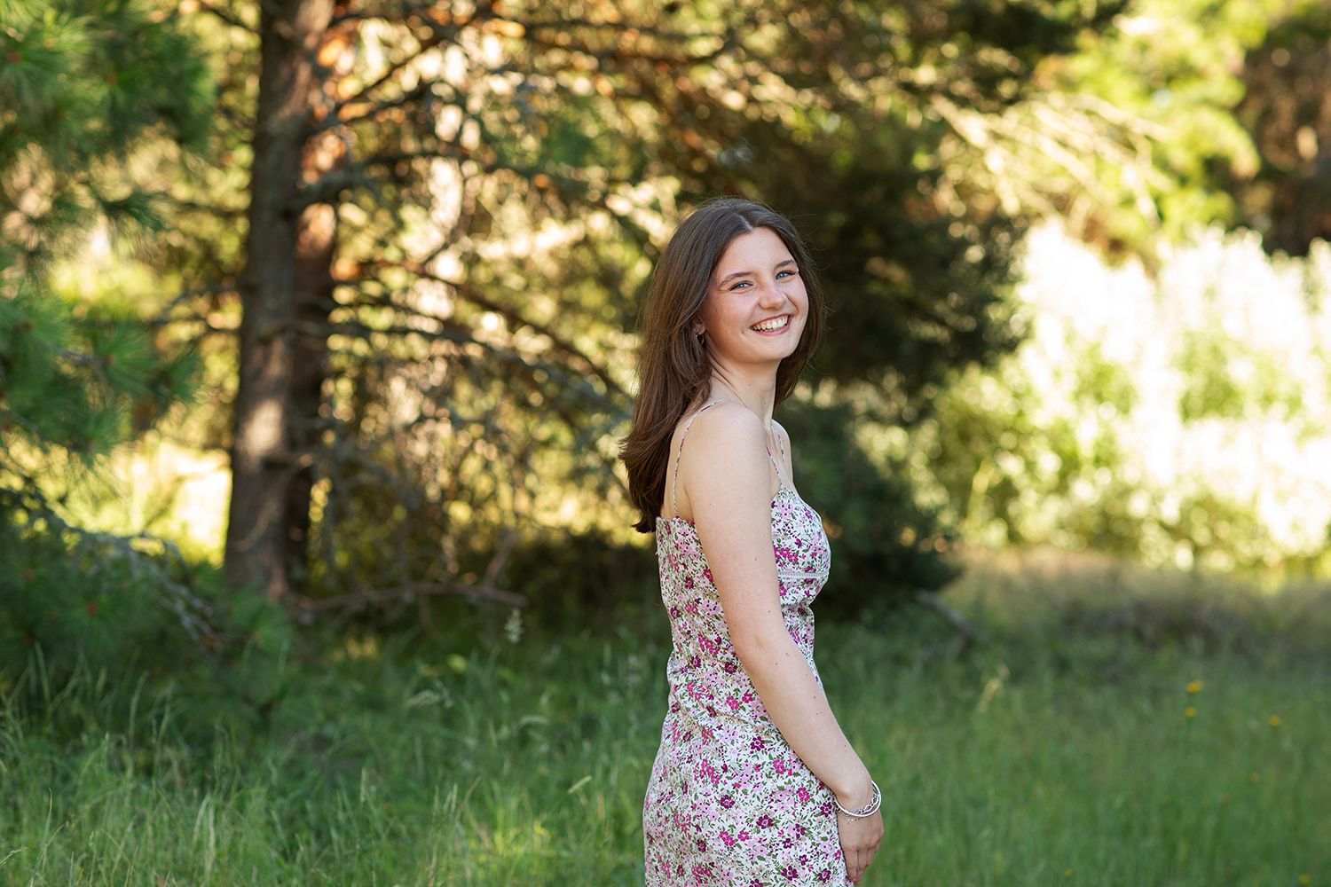 Young woman in floral dress smiling outdoors in a sunny green forest setting.