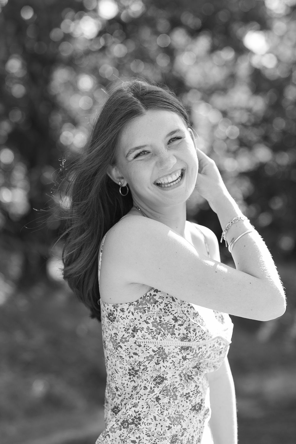 Black and white portrait of a smiling young woman in a floral dress outdoors, hair blowing in the breeze.