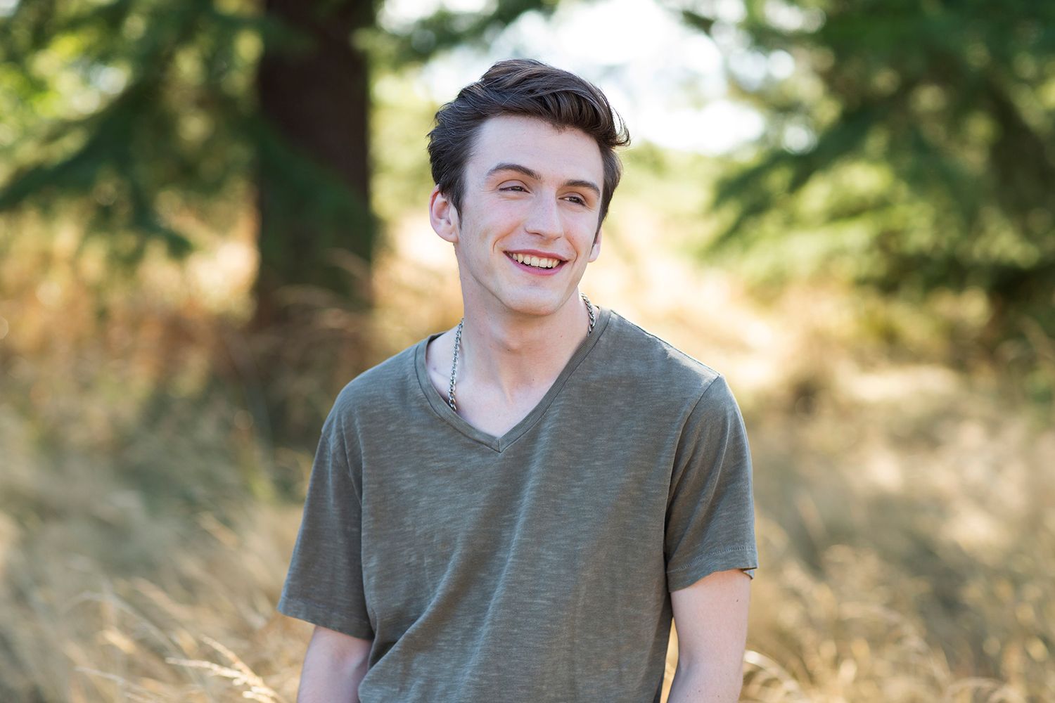 Smiling young man in gray t-shirt standing outdoors in a sunny field with trees in the background.