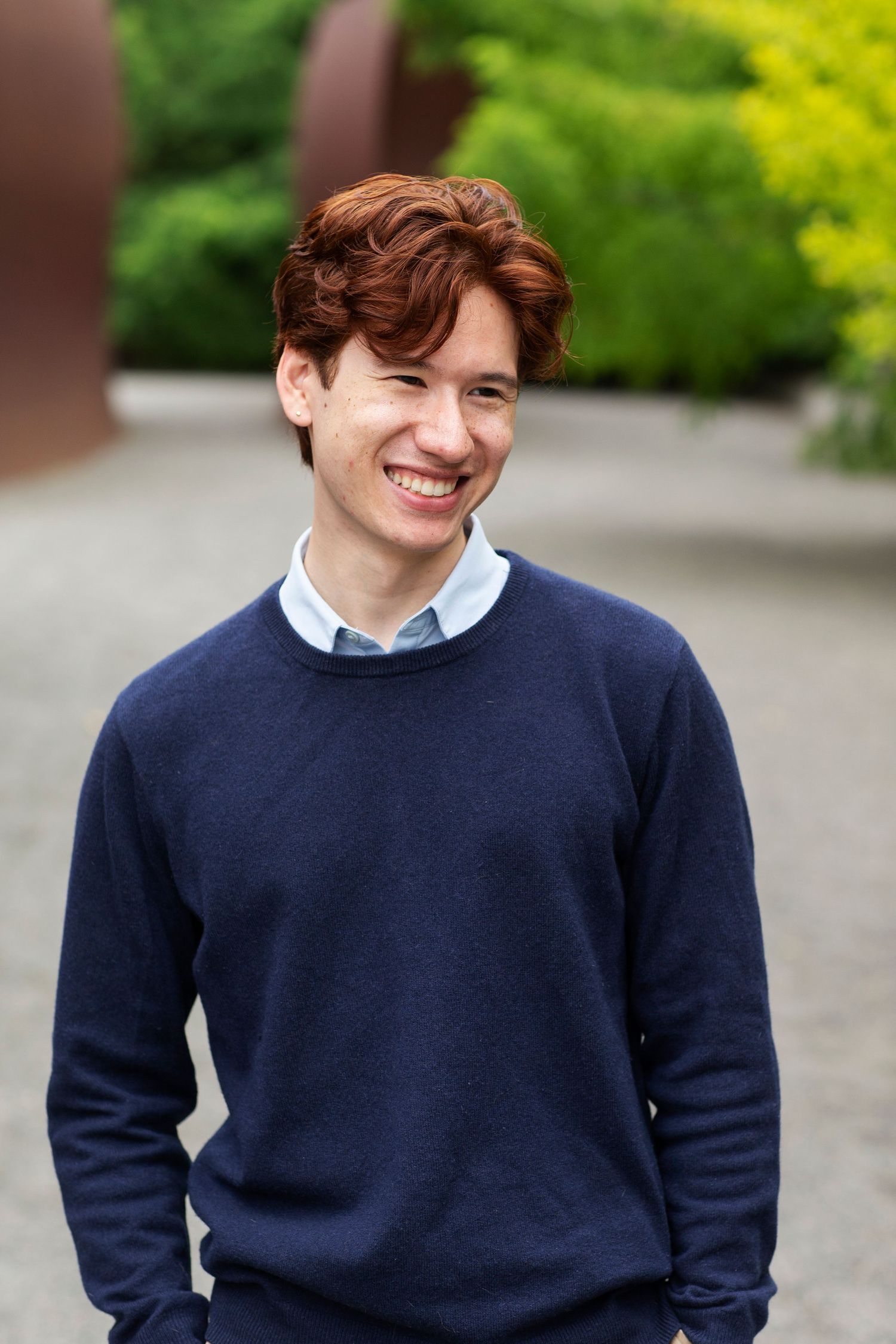 Young man in navy sweater smiling outdoors in garden setting.