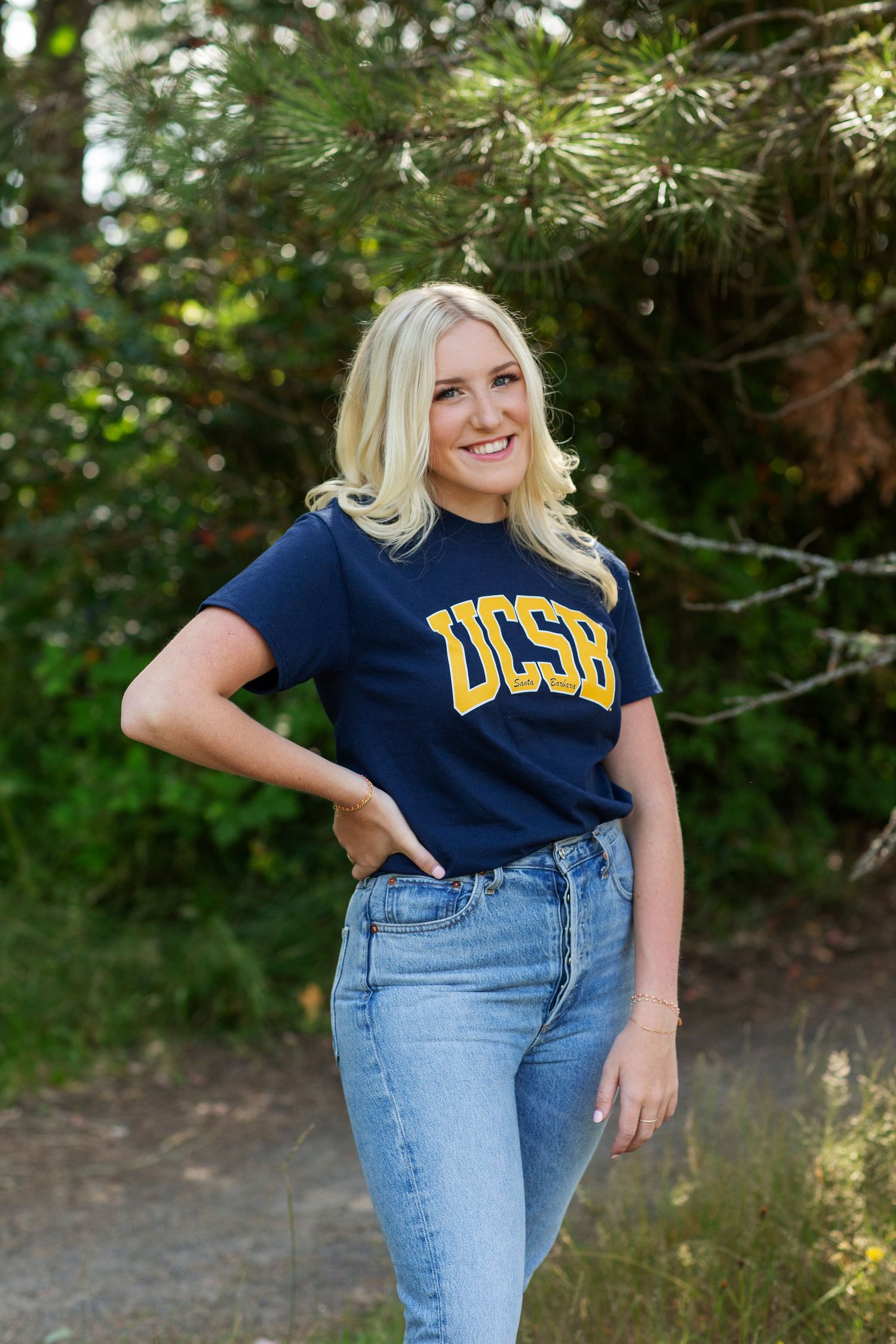 Blonde woman wearing a navy UCSB college t-shirt and jeans posing outdoors near pine trees.