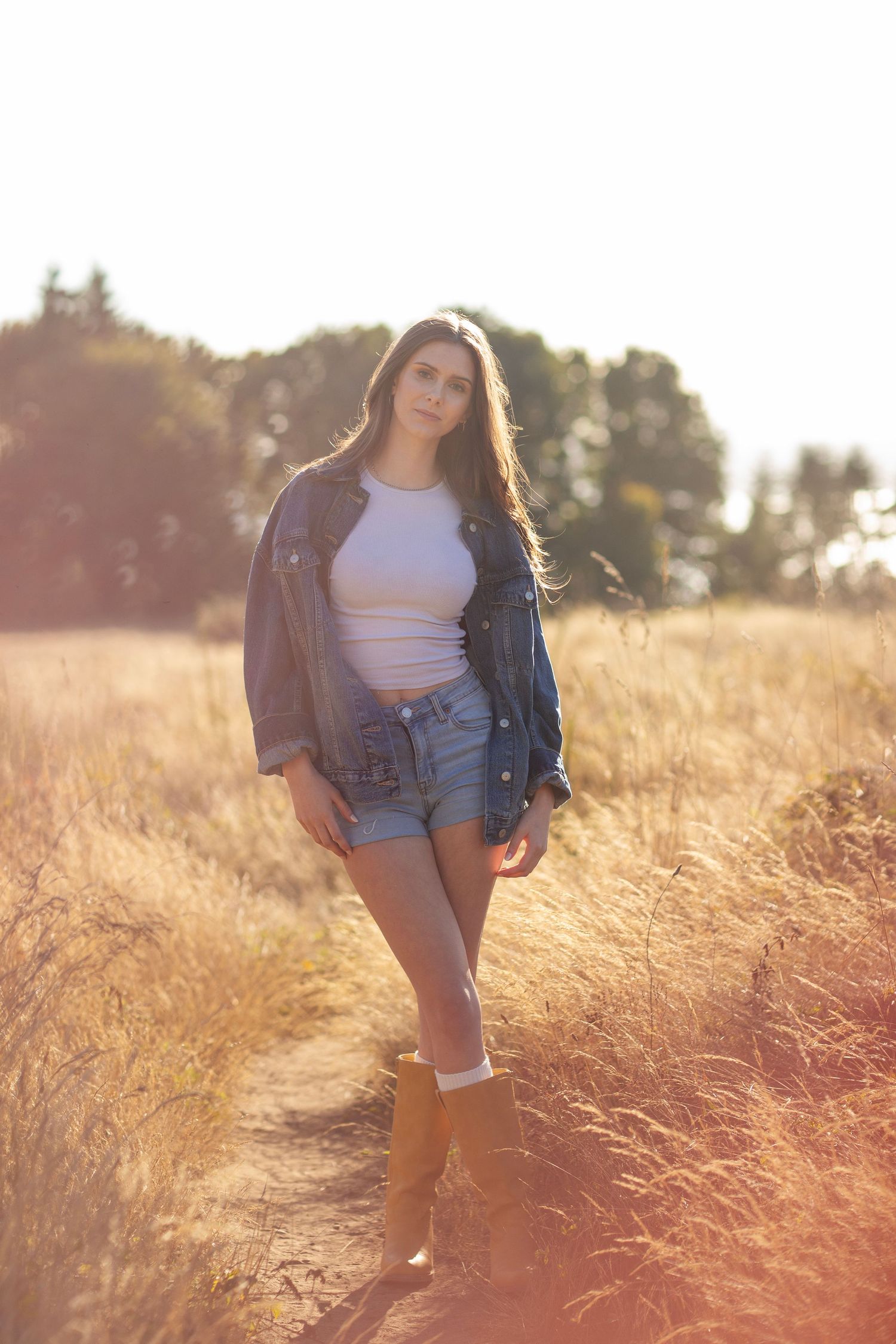 Young woman in denim jacket and shorts posing confidently on a golden sunlit field trail.