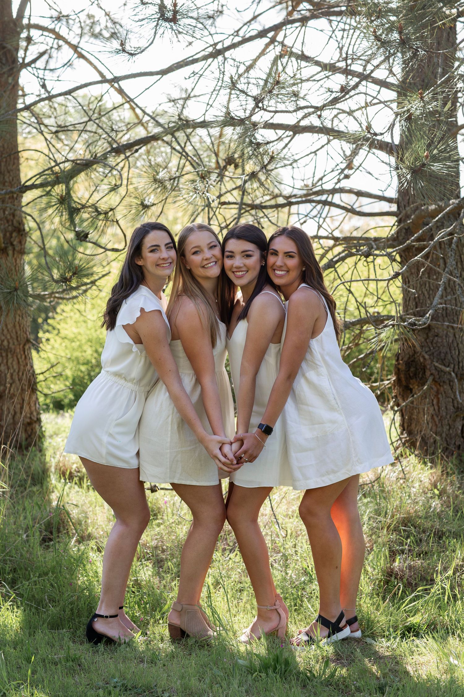 Four young women in matching white dresses pose together outdoors among pine trees on a sunny day.