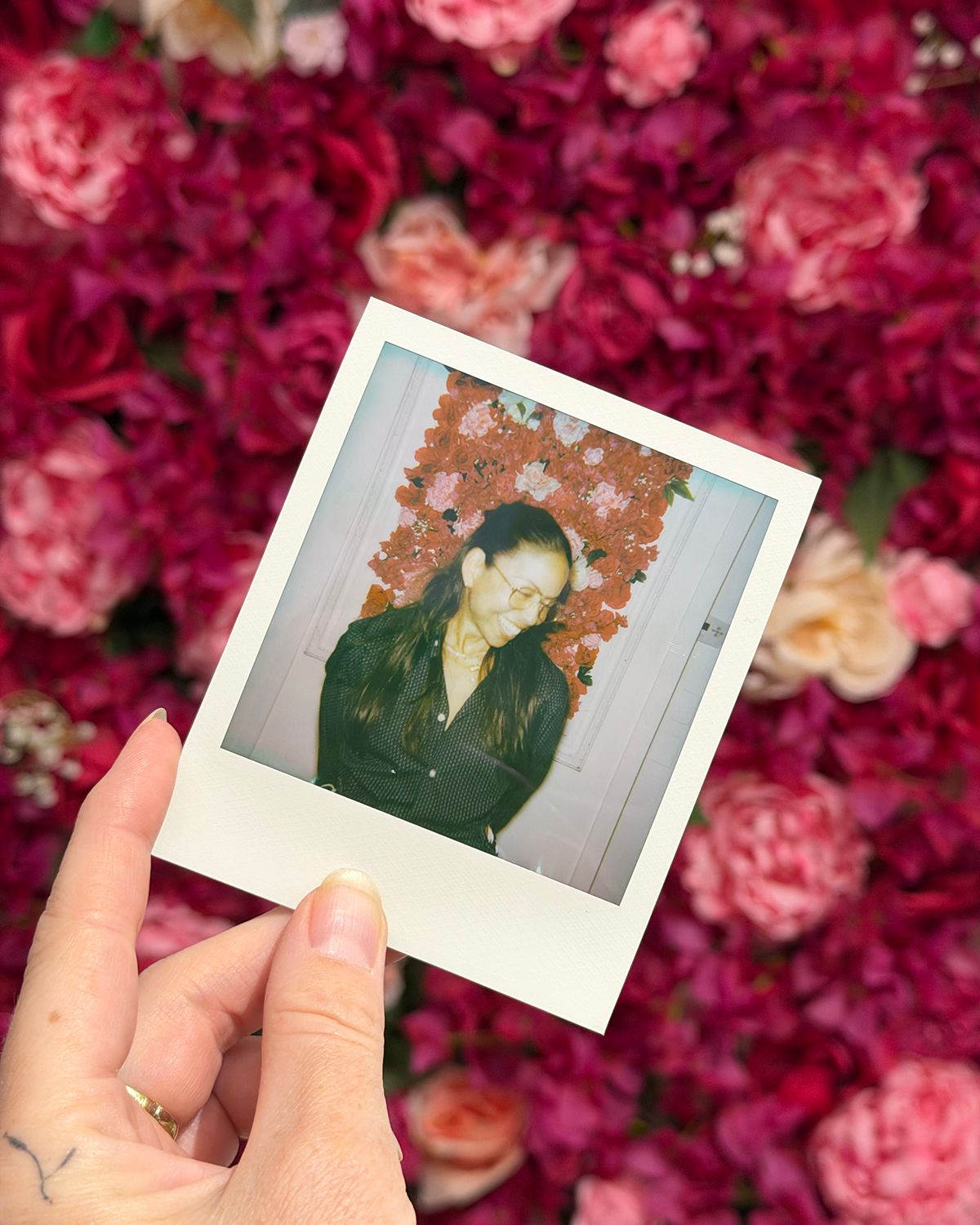 A hand holds a Polaroid photo of a person in black against a vibrant pink and red floral wall backdrop.