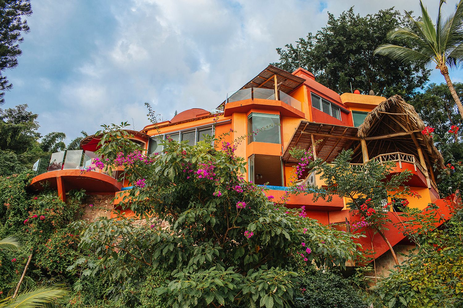 Vibrant orange multi-level tropical home surrounded by lush greenery and colorful bougainvillea flowers.