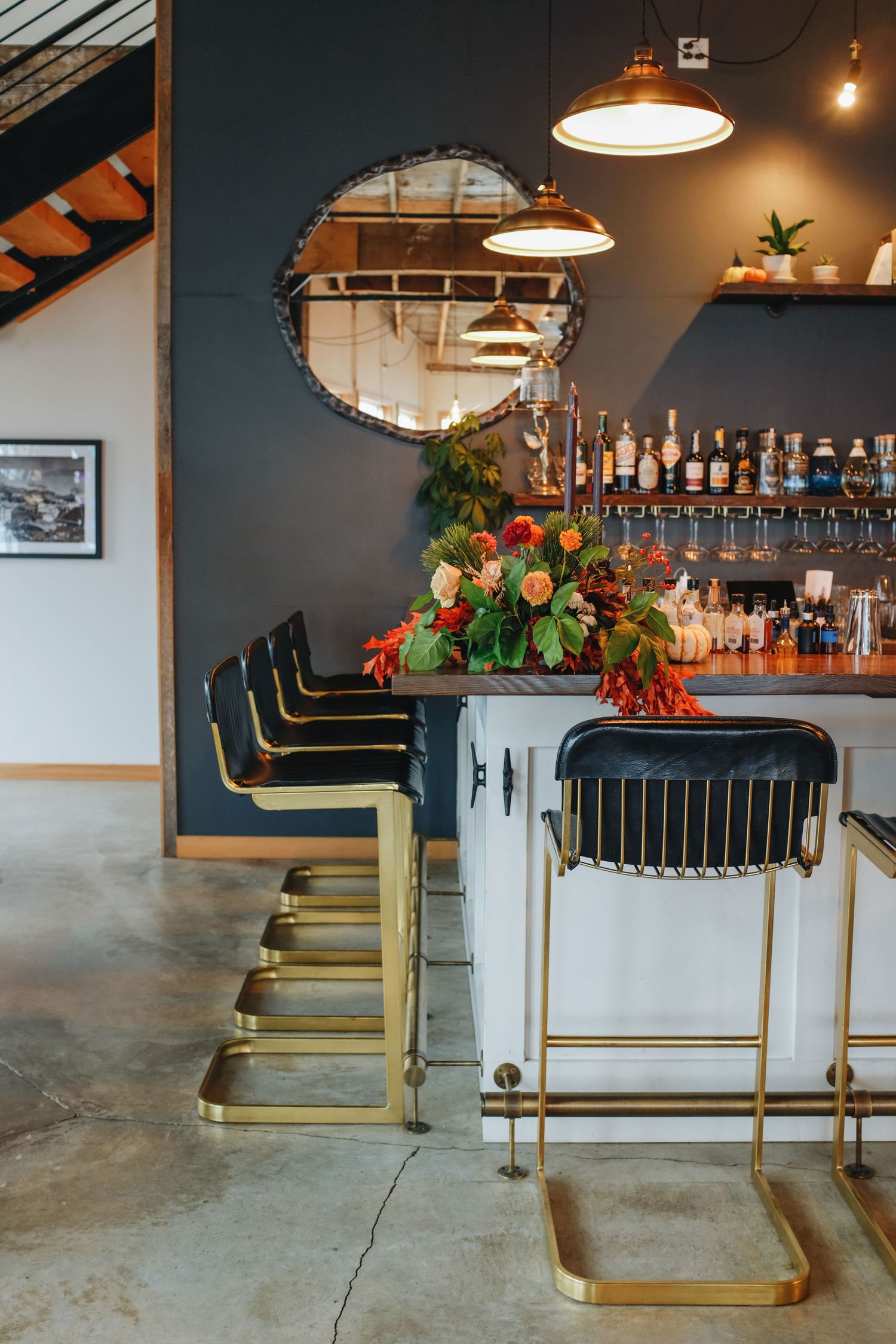 Stylish bar area with gold bar stools, floral arrangement, pendant lights, and shelves of bottles against a dark blue wall.