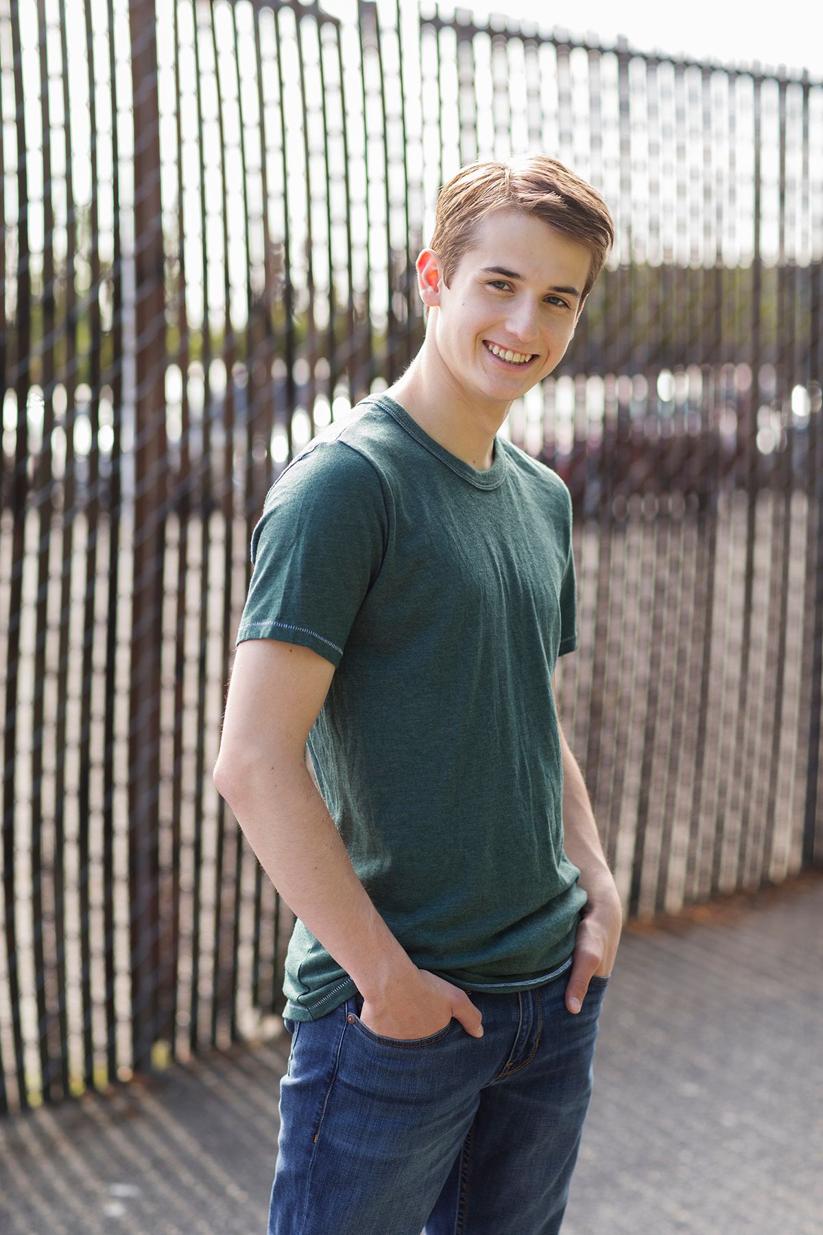 Smiling young man in green t-shirt and jeans standing in front of metal fence outdoors.