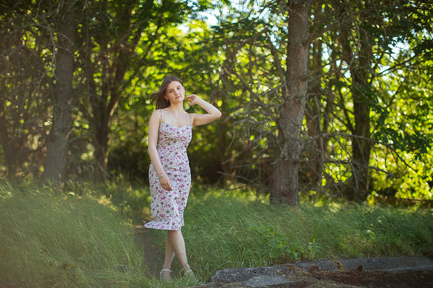 Young woman in floral sundress standing among lush green trees in a sunlit forest setting.