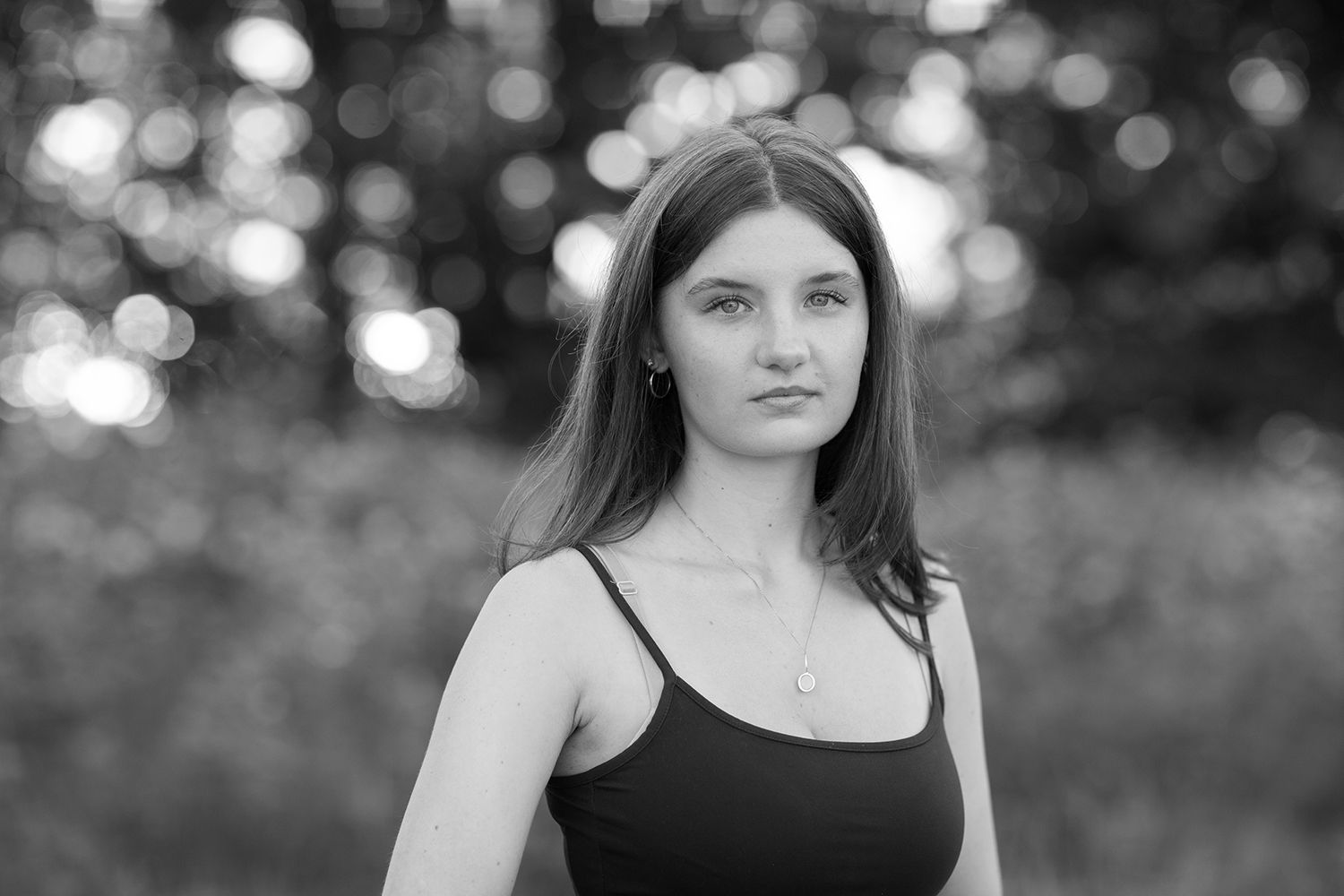 Black and white portrait of a young woman in a tank top with a necklace, posing outdoors with bokeh background.