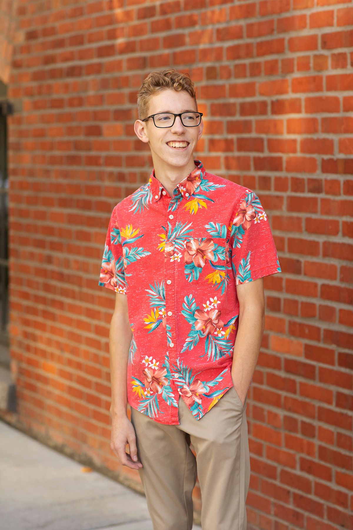 Smiling teenage boy wearing red floral Hawaiian shirt and glasses standing against a brick wall outdoors.