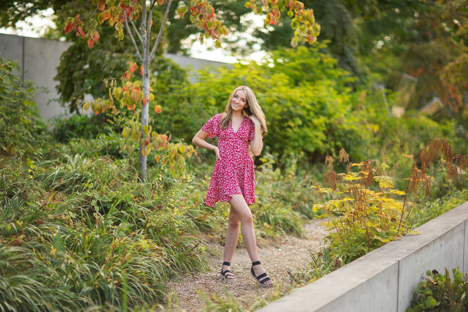 Blonde woman in red floral dress posing in a vibrant autumn garden setting.