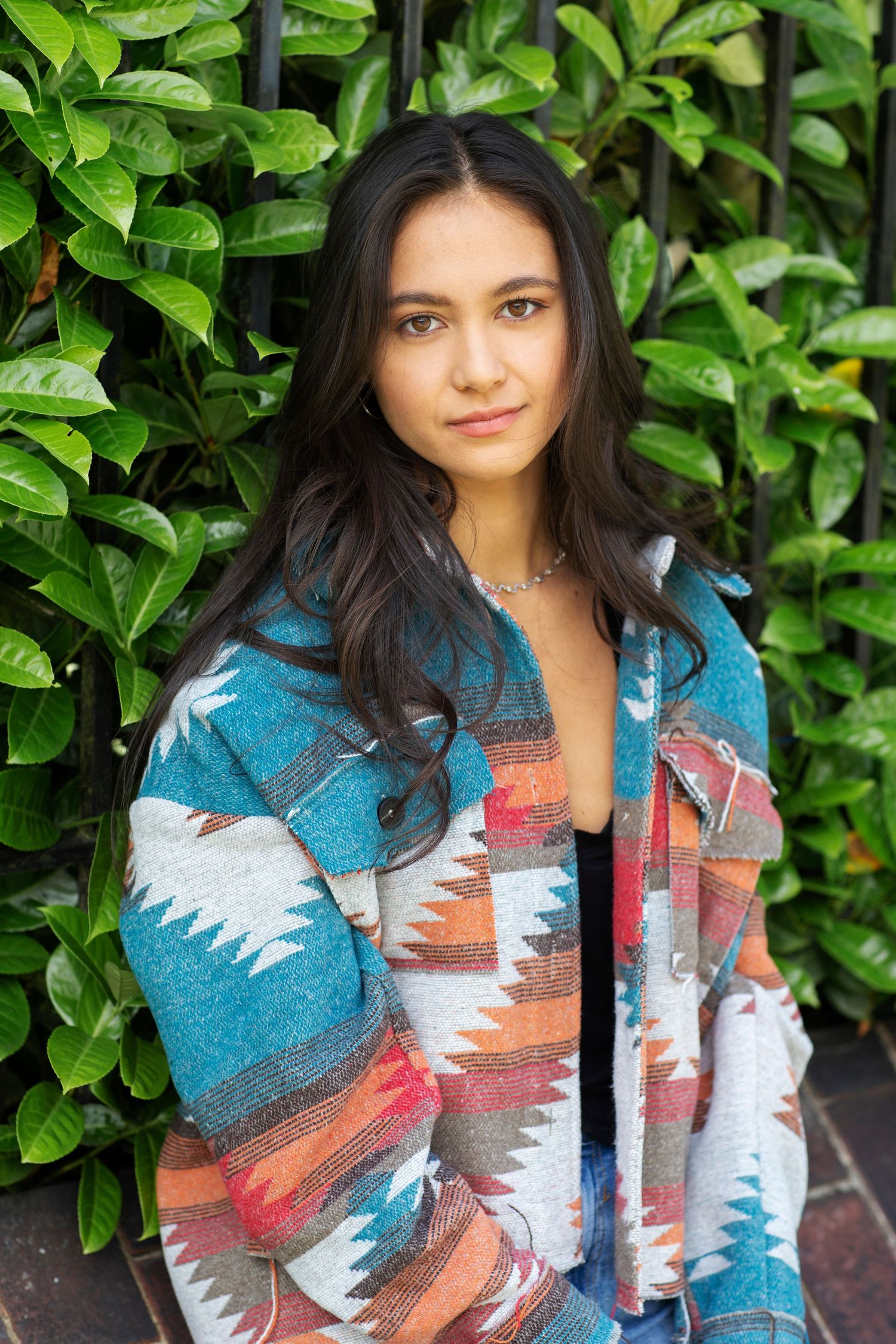 Young woman in teal aztec print jacket posing seriously against a lush green leaf wall.