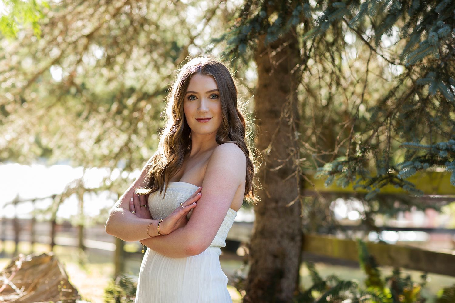 Young woman in white dress with arms crossed smiling outdoors near trees and wooden fence.