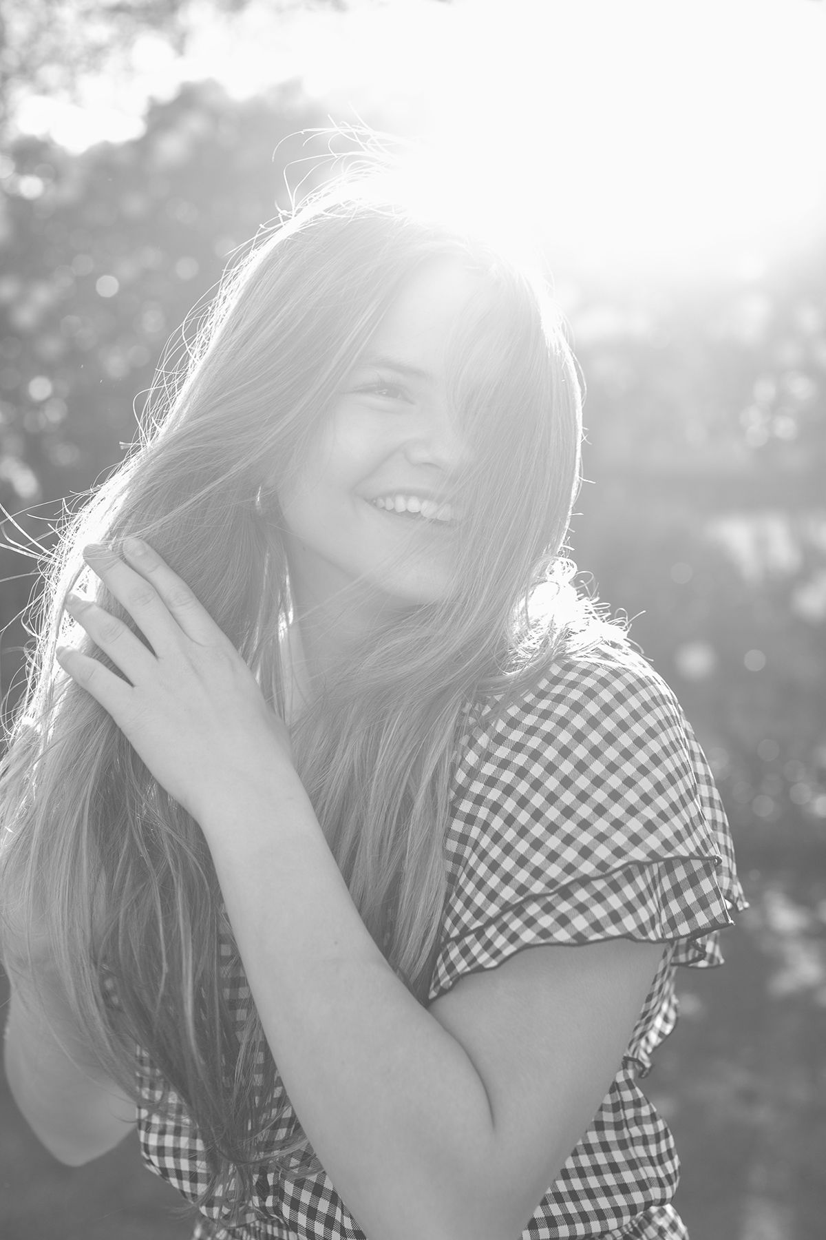 Black and white portrait of a smiling young woman with long hair in a gingham top, bathed in bright sunlight outdoors.