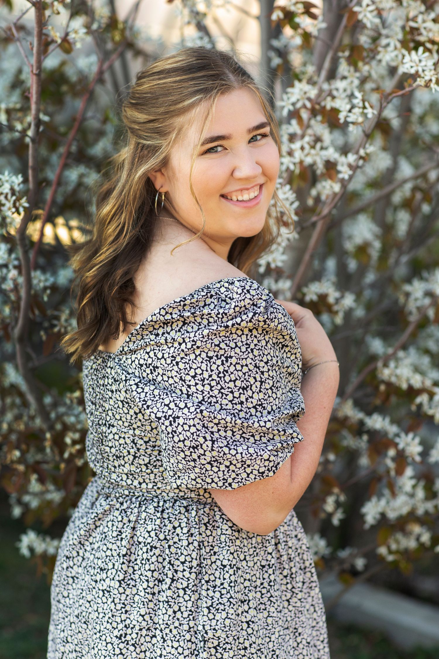 Young woman in floral off-shoulder dress smiles outdoors near blooming white flowering shrubs in natural light.