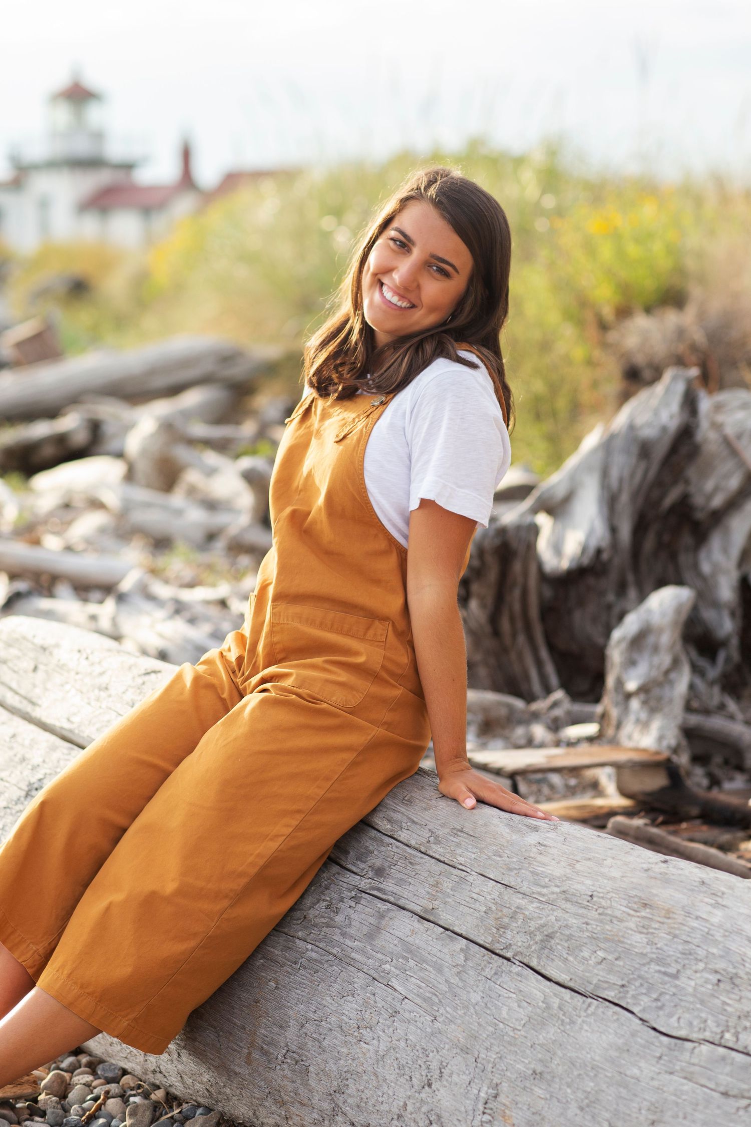 Young woman in mustard overalls sitting on driftwood log at a scenic beach with lighthouse in background.