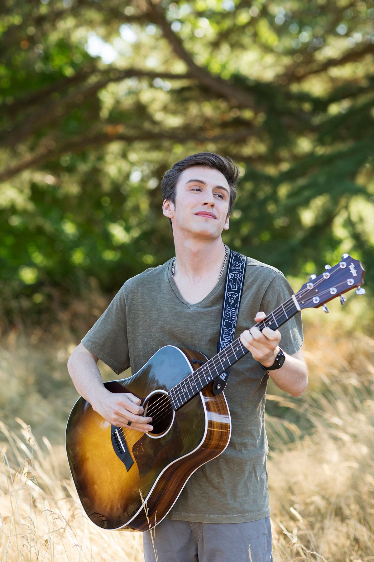 Young man smiling while playing acoustic guitar outdoors in a sunny field with green trees in the background.