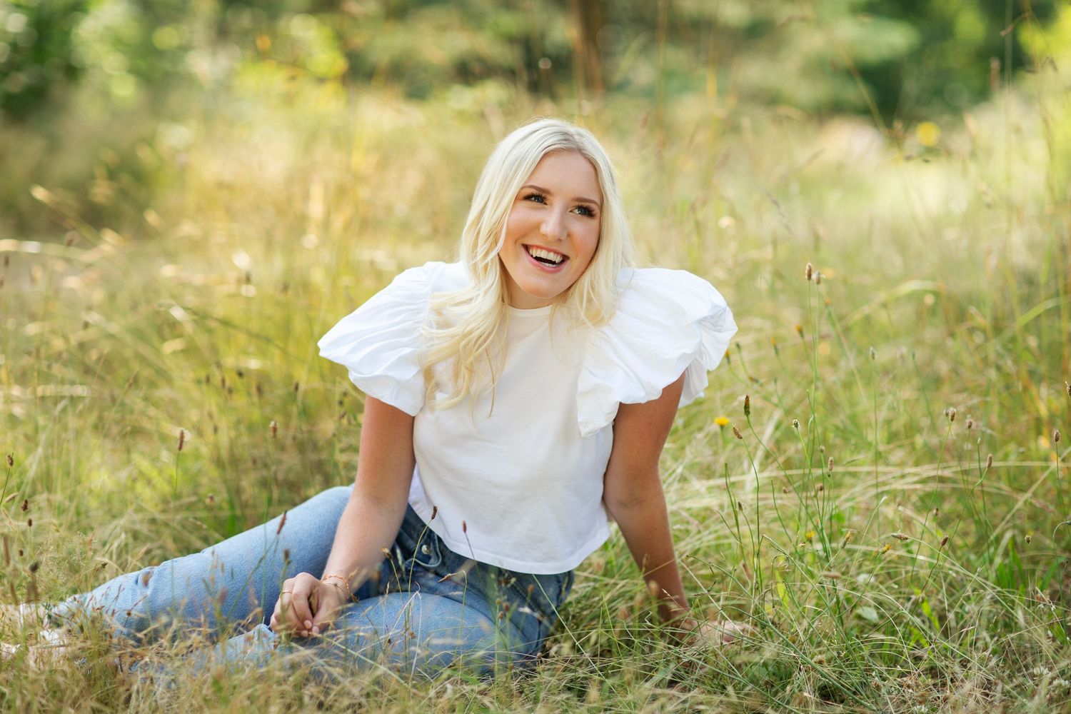 Happy blonde woman sitting in a sunny meadow wearing a white ruffle top and blue jeans, smiling.