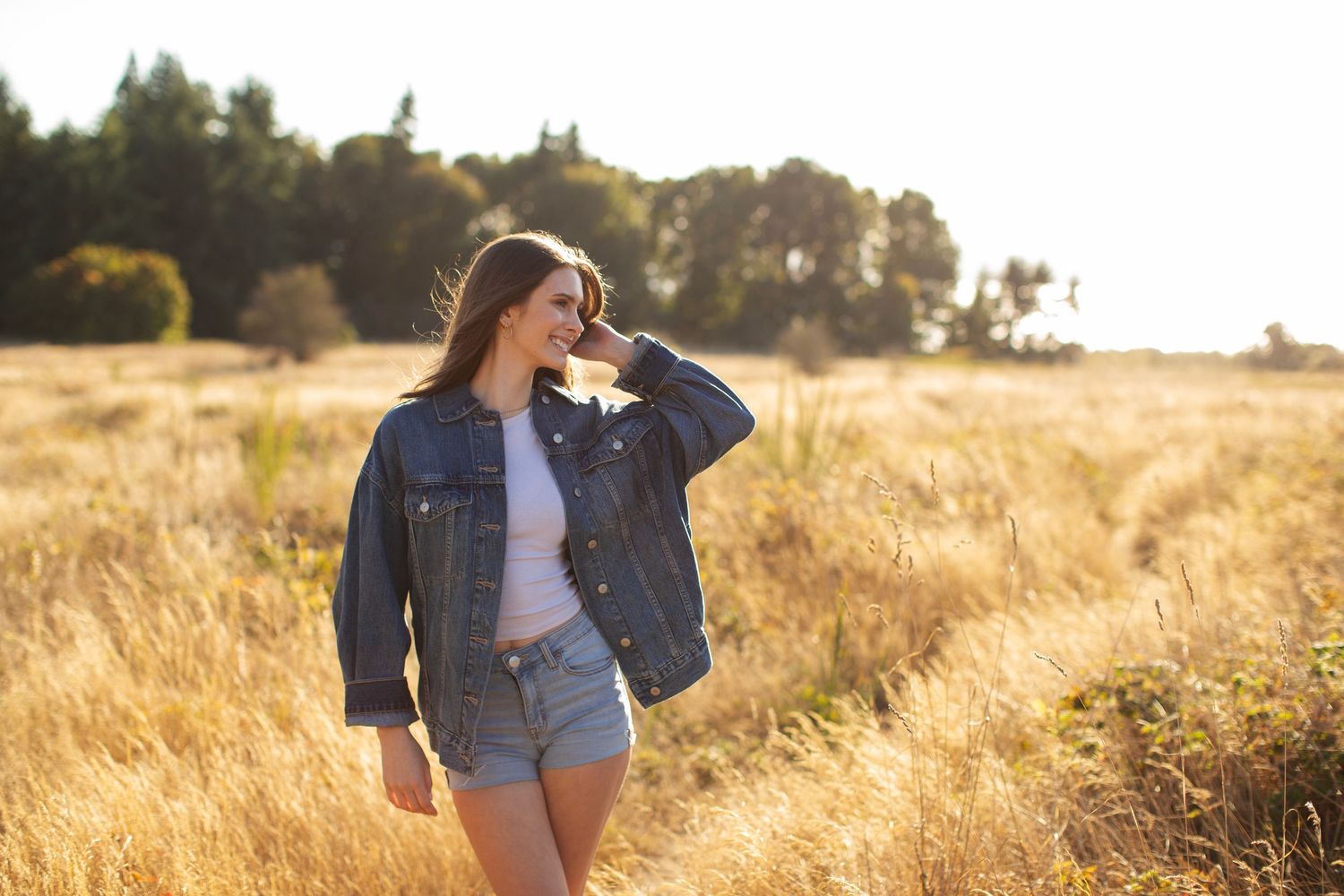 Young woman in oversized denim jacket and shorts smiling in a sun-drenched field.