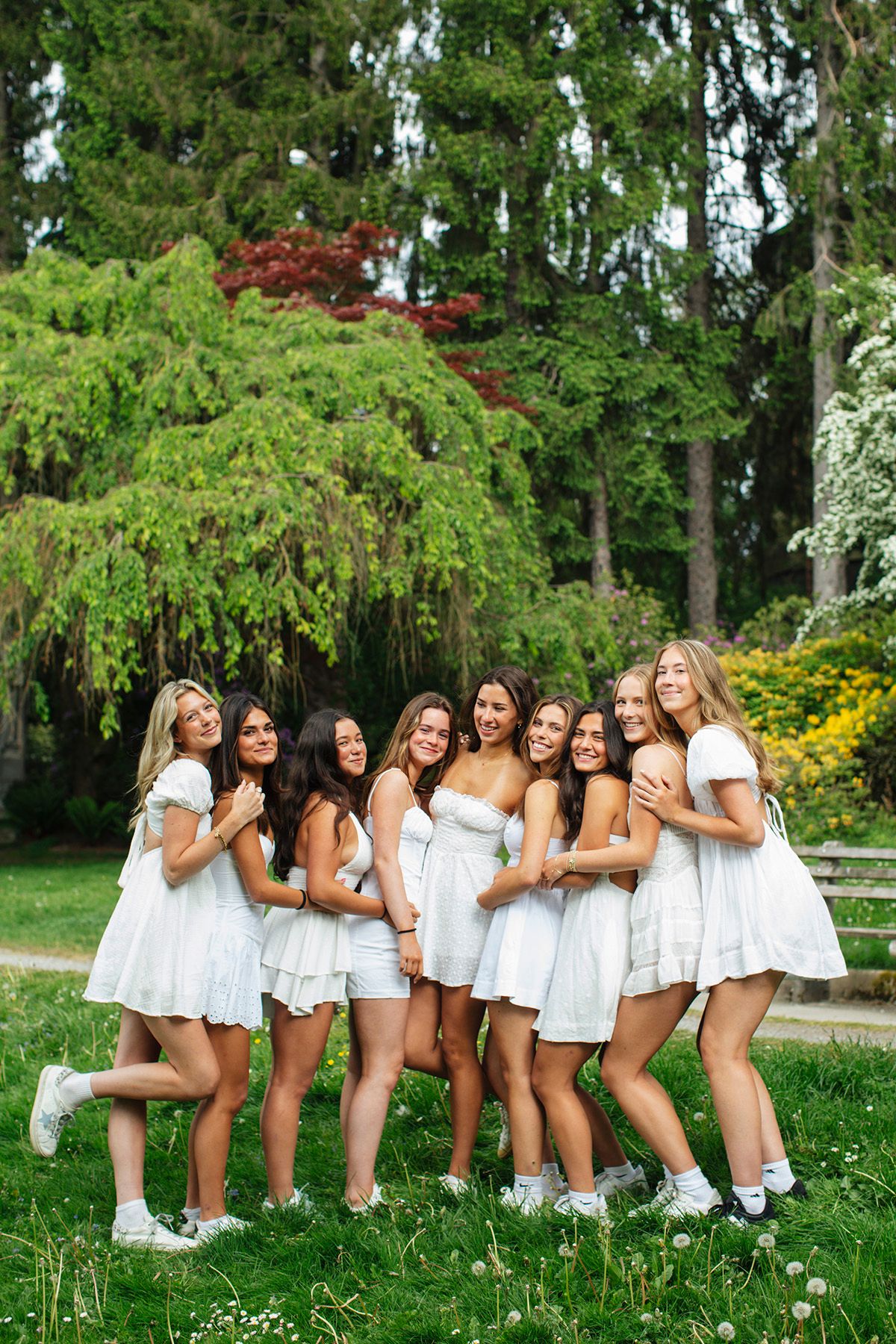 Group of young women in matching white dresses smiling and hugging outdoors in a lush green park.