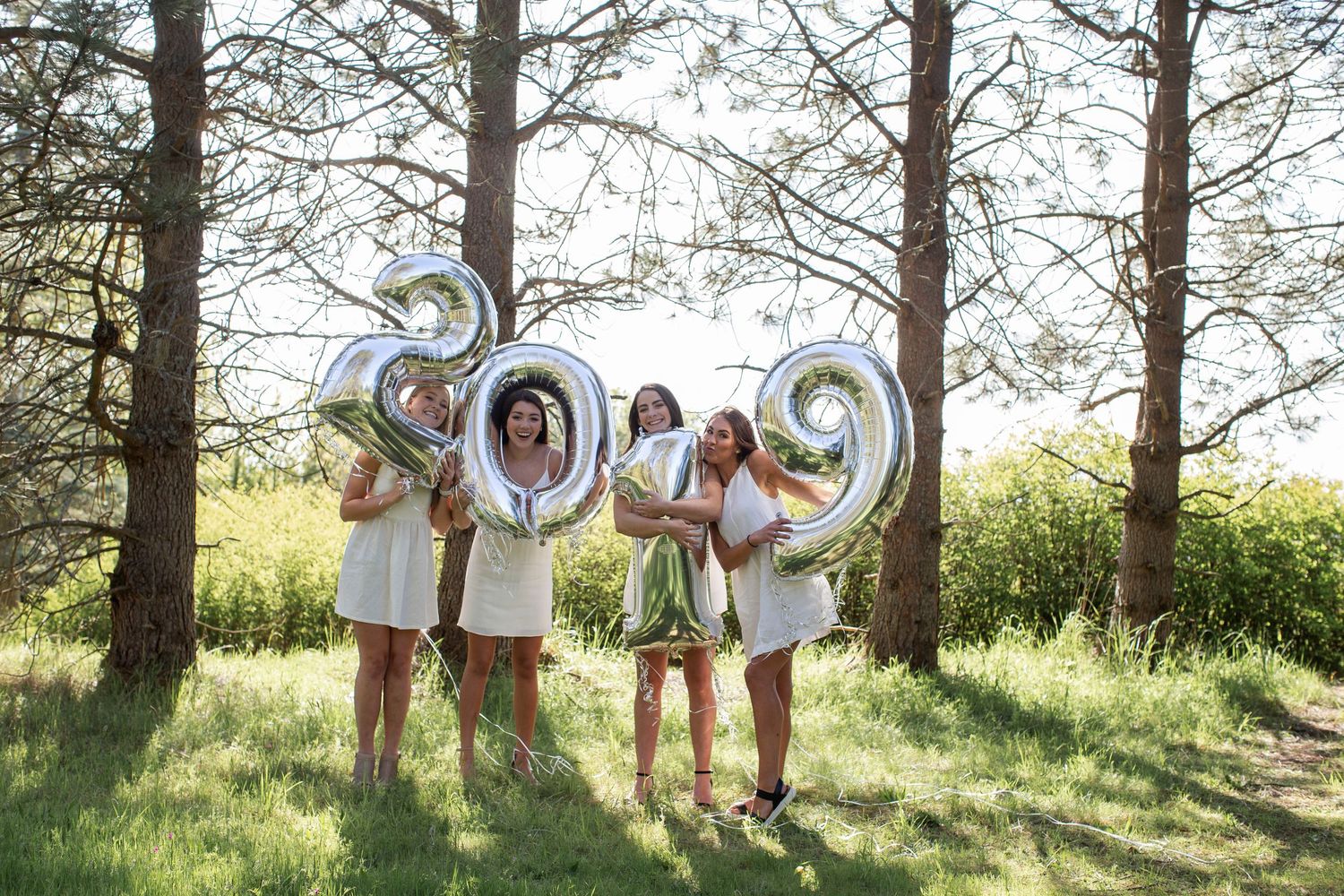 Four women in white dresses hold silver 2019 balloon numbers outdoors among trees on a sunny day.