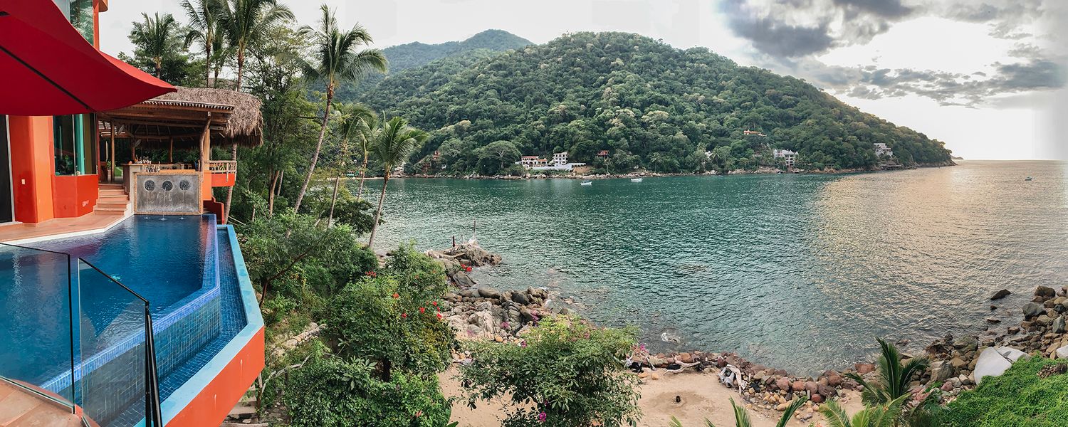 Panoramic view of colorful tropical villa with pool overlooking scenic bay and lush mountains.