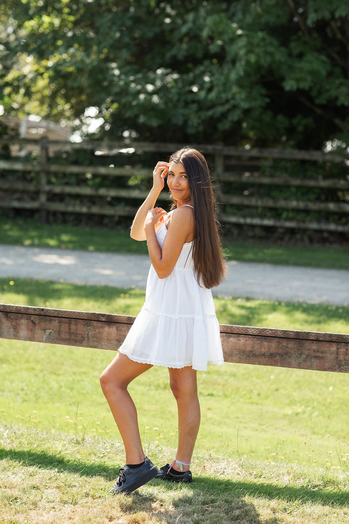 Young girl in white sundress and black shoes poses on green grass in sunny outdoor park setting.