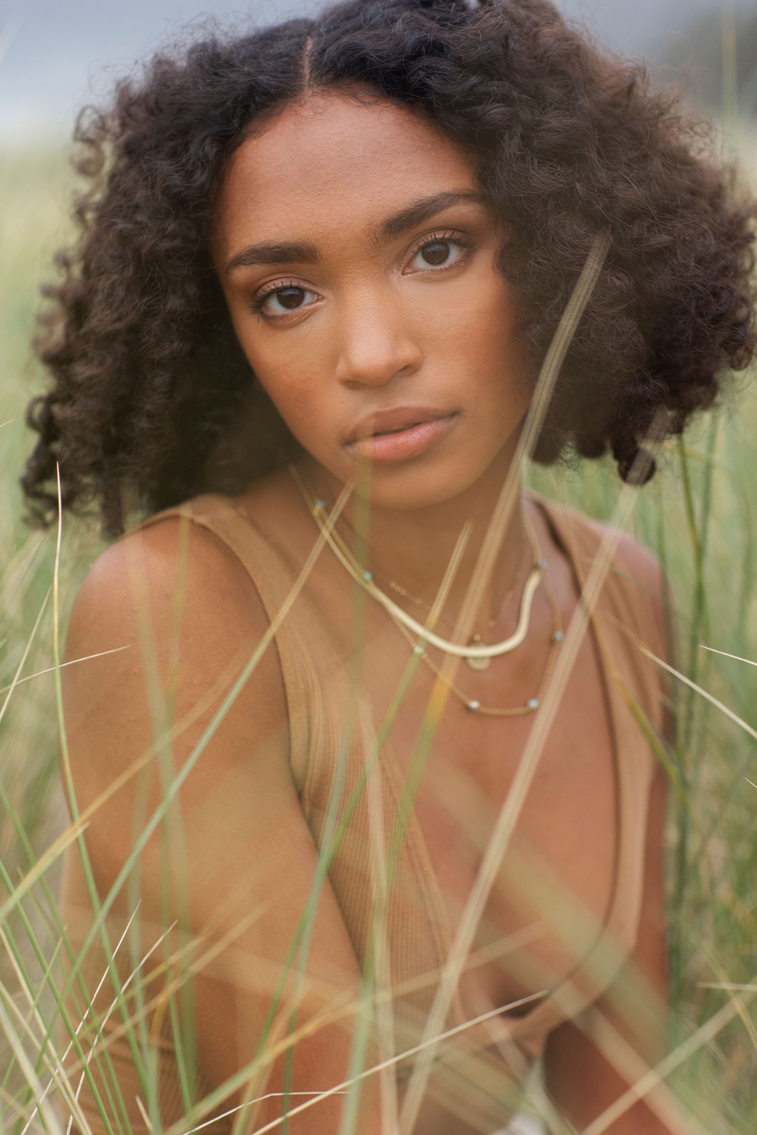 Young woman with curly hair wearing a tan top and gold necklaces sitting in tall grass outdoors.