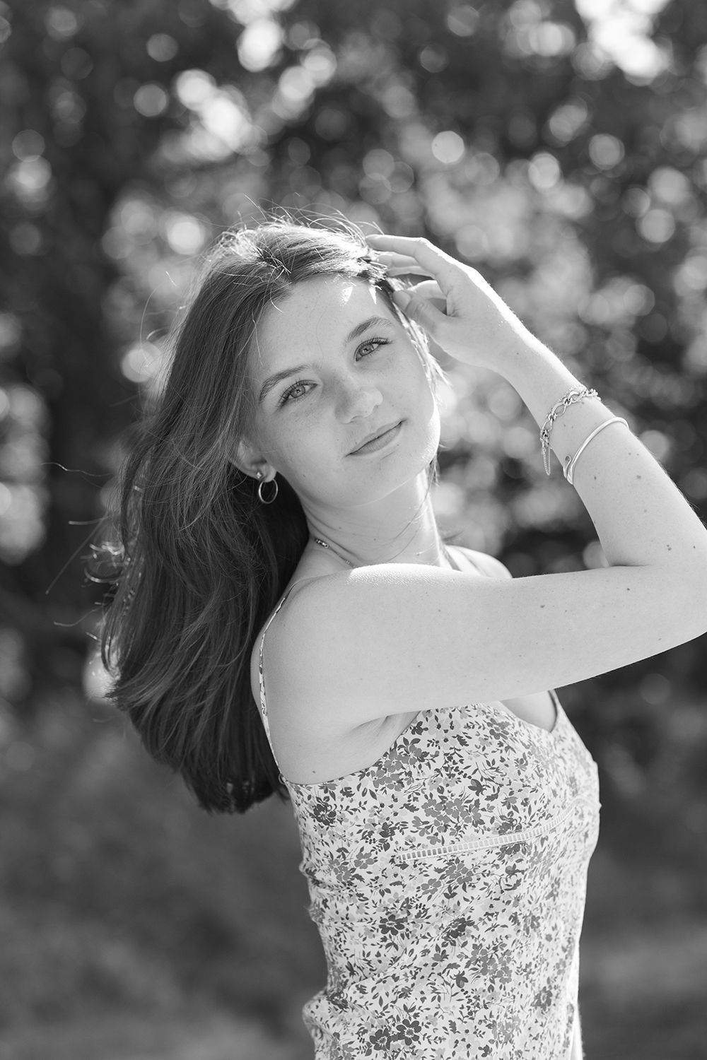 Black and white portrait of a young woman in a floral dress, hand in hair, posing outdoors with blurred trees.