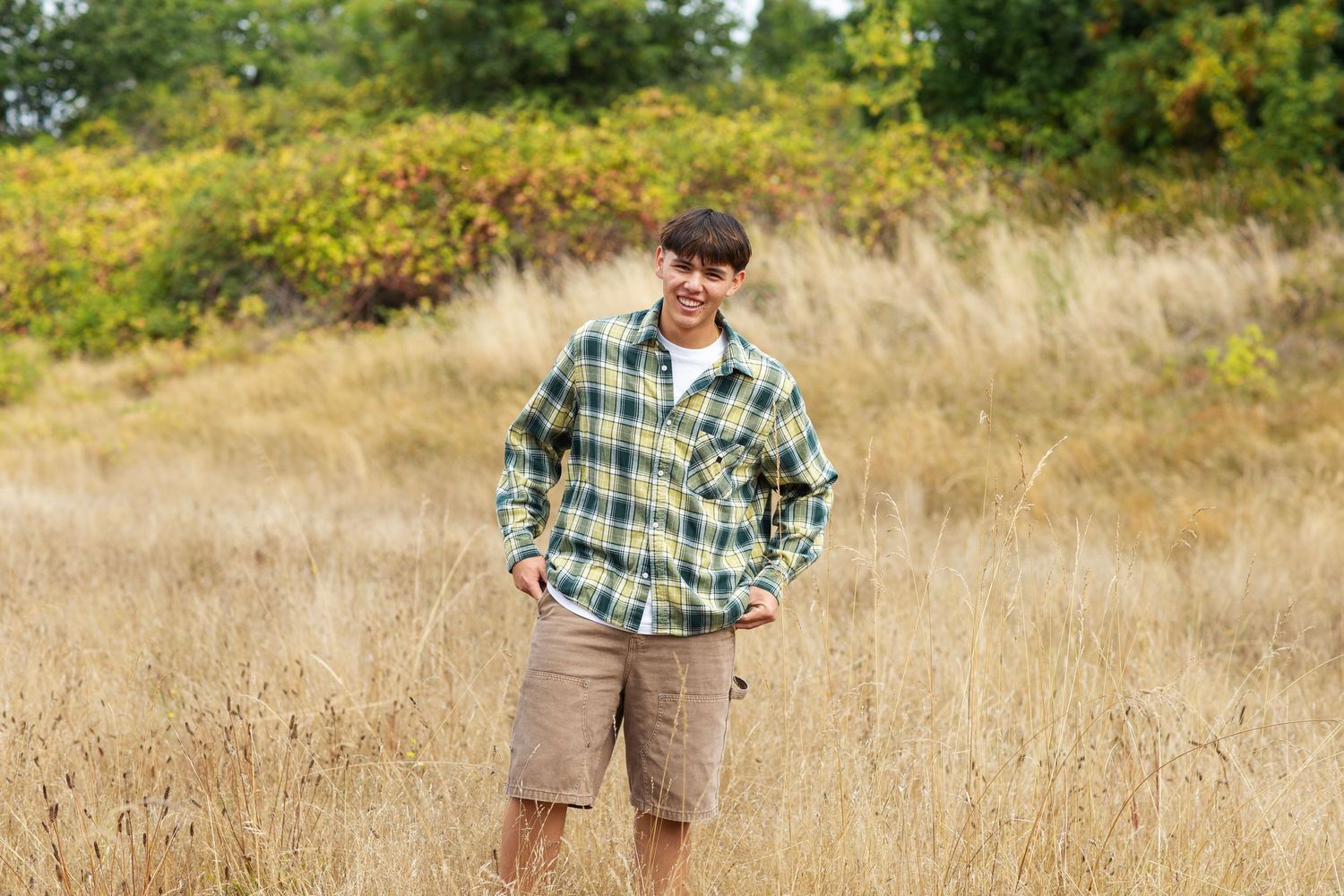 Teen boy in green plaid flannel shirt and shorts posing in tall dry grass field.