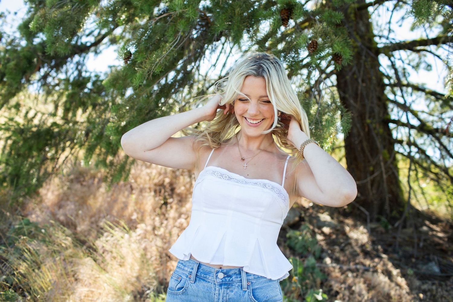 Laughing blonde woman in white ruffled cami top and jeans with hands in hair near pine trees outdoors.