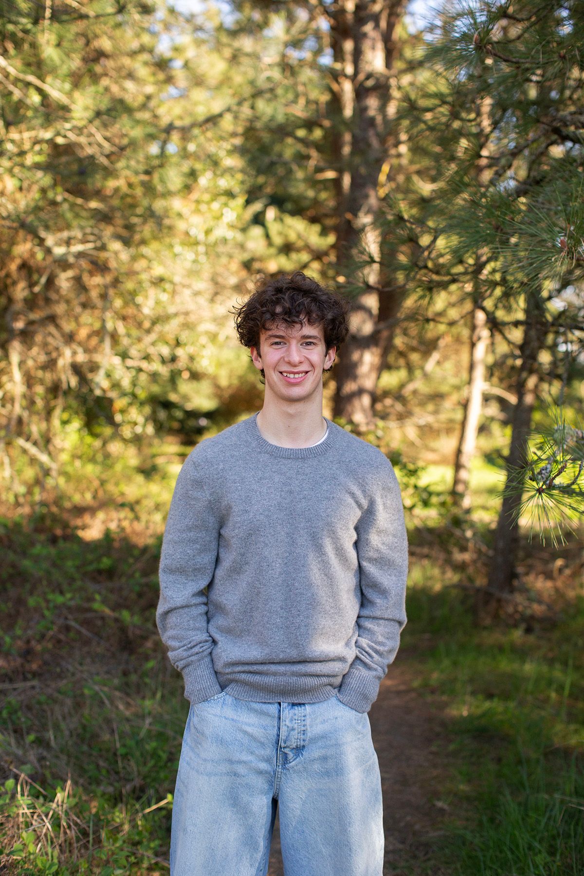 Young man with curly hair smiling outdoors in a gray sweater and jeans, standing in a sunlit forest path.